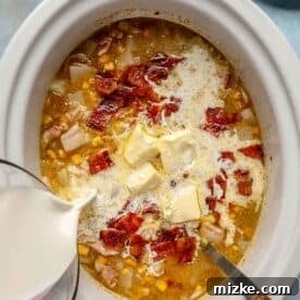 Cubes of unsalted butter and a stream of creamy half and half are being added to the simmering corn chowder inside the slow cooker insert.