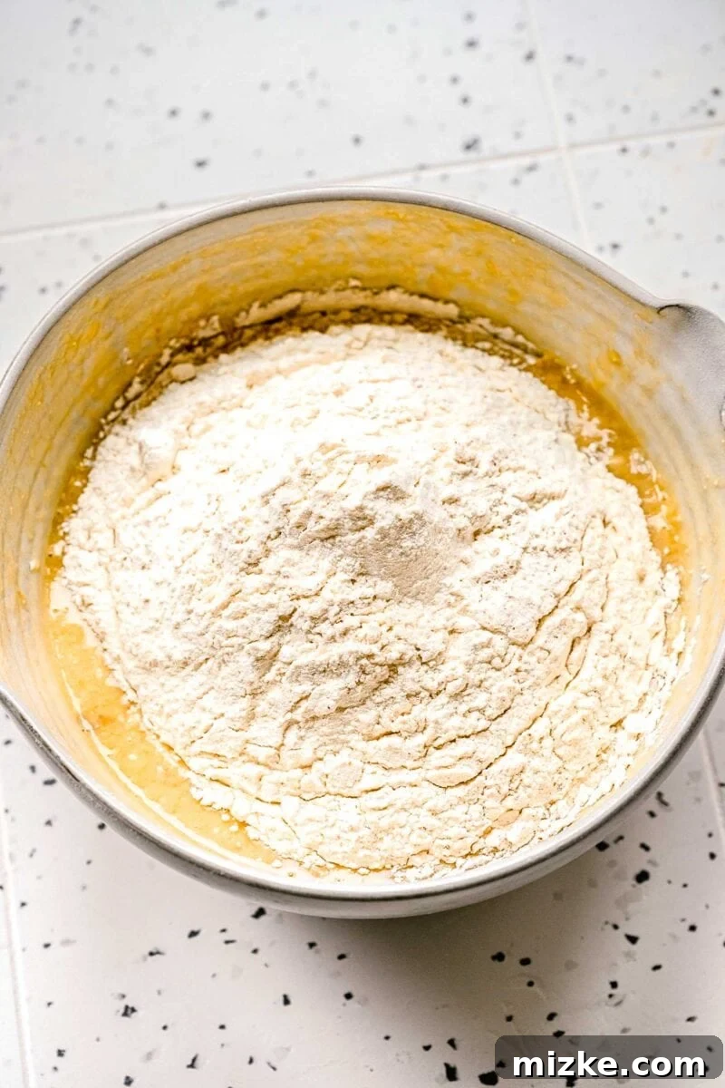 Sift dry ingredients directly into the wet for a lump-free banana blueberry muffin batter. Dry ingredients, including flour, baking powder, baking soda, salt, cinnamon, and nutmeg, being added to the wet banana blueberry muffin batter.
