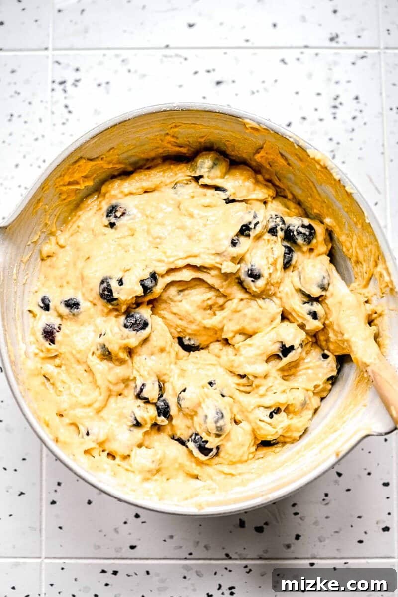 A perfectly combined banana blueberry muffin batter, ready for baking. Fully mixed banana blueberry muffin batter in a bowl with a wooden spoon, ready for portioning into muffin tins.