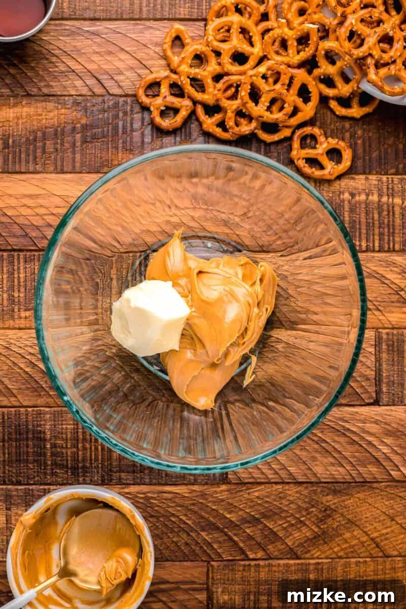 Creamy peanut butter and softened butter being combined in a clear bowl next to a pile of mini pretzels, illustrating the start of the filling process