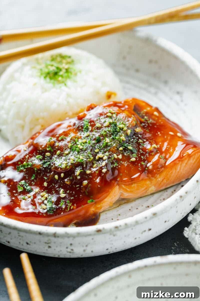 Perfect Air Fryer Teriyaki Salmon 3 Close-up image of a perfectly cooked teriyaki glazed salmon fillet, topped with furikake, served on a ceramic plate next to fluffy white rice and wooden chopsticks.