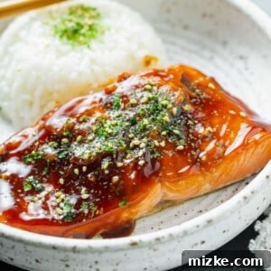 Perfect Air Fryer Teriyaki Salmon 10 Close-up image of teriyaki glazed salmon on a speckled ceramic plate with furikake sprinkled on top, next to a mound of white rice with wooden chopsticks.