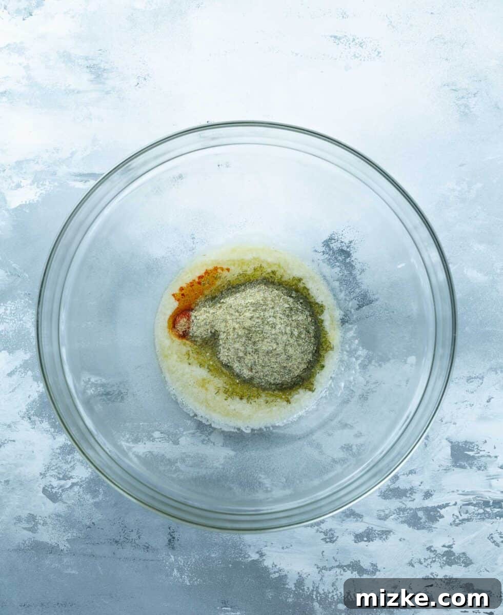 Melted butter in a clear glass bowl with ranch seasoning, garlic powder, and smoked paprika sprinkled on top, ready for mixing.