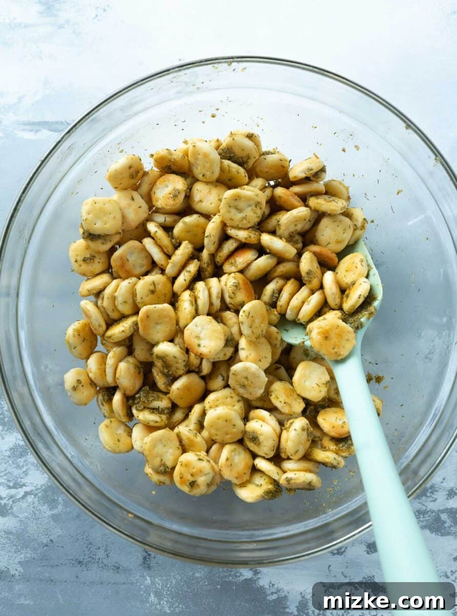 Oyster crackers being tossed in a large clear bowl with a mint-colored spatula, thoroughly coated in the butter and seasoning mixture.
