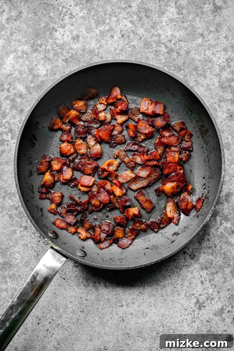 Golden Bacon Roasted Brussels Sprouts 4 Strips of bacon sizzling and crisping up in a grey nonstick skillet on the stovetop.