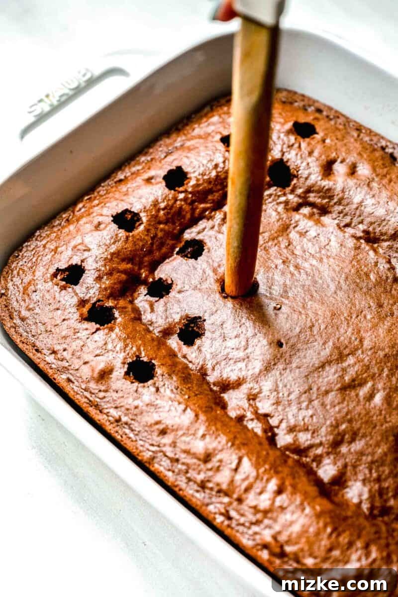 Decadent Chocolate Pudding Poke Cake 7 The end of a wooden spoon gently poking holes into the surface of the freshly baked chocolate cake, still in its white baking dish.