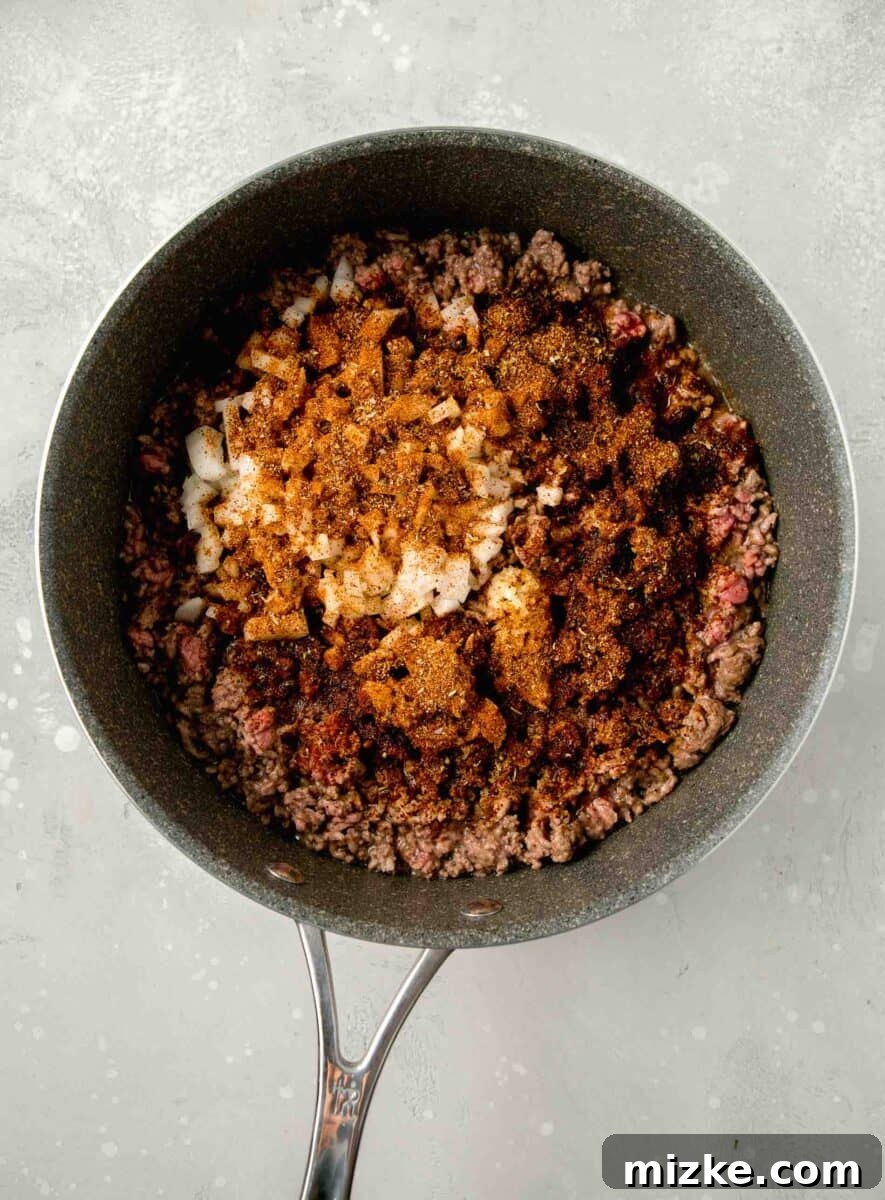 Step 2: Adding aromatics and seasoning to the beef. Diced onions, minced garlic, and taco seasoning being added to the browned ground beef in the skillet, ready to be stirred.