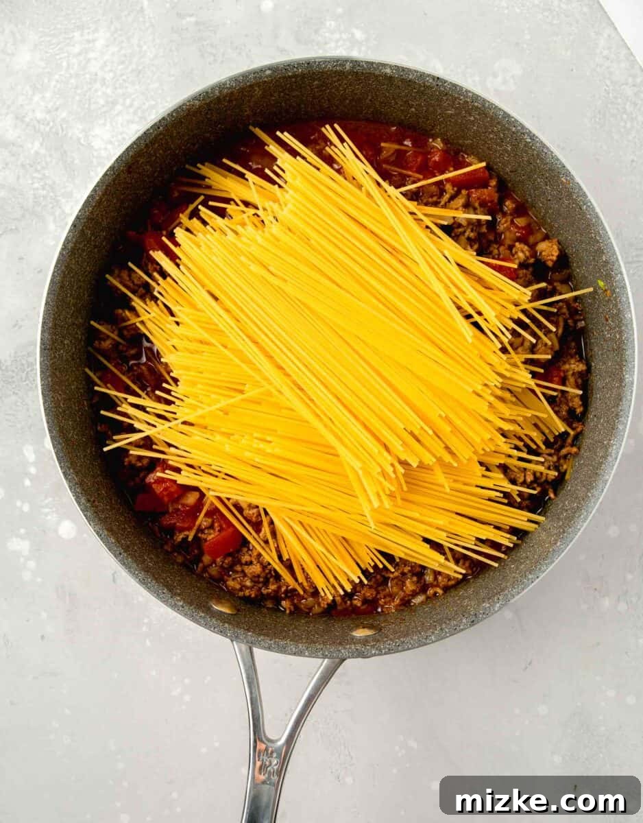 Step 4: Incorporating the spaghetti pasta. Uncooked thin spaghetti, broken in half, being added to the skillet with the ground beef and sauce mixture, ensuring it's submerged.