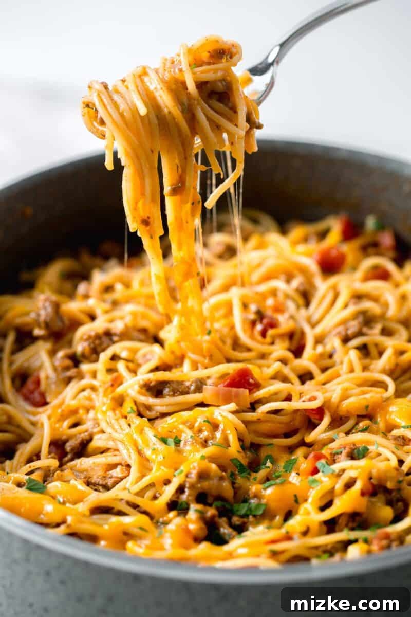 Enjoying a cheesy serving of one-pot taco spaghetti. A fork lifting a portion of cheesy taco spaghetti out of a skillet, with a visible cheese pull.