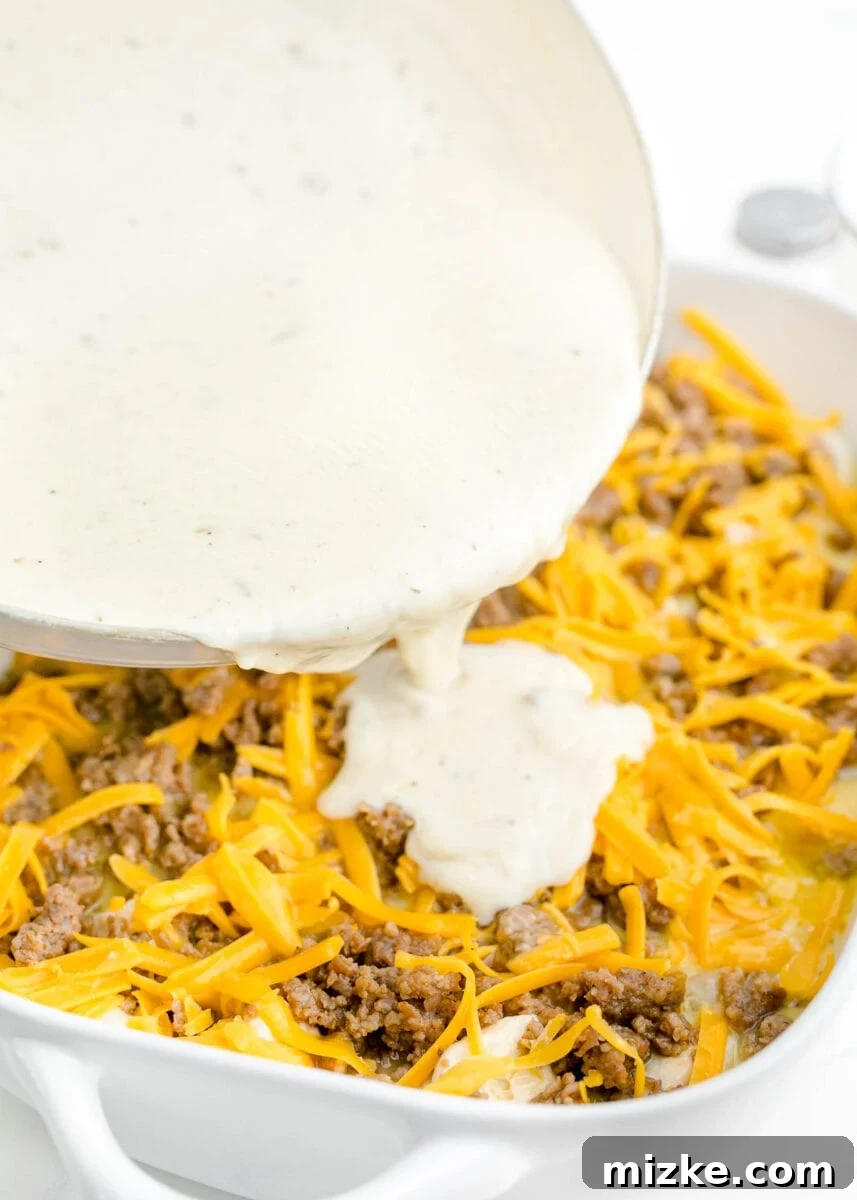 Southern Comfort Biscuit Bake 11 thickened white gravy being poured on top of baking dish with browned sausage and shredded cheese