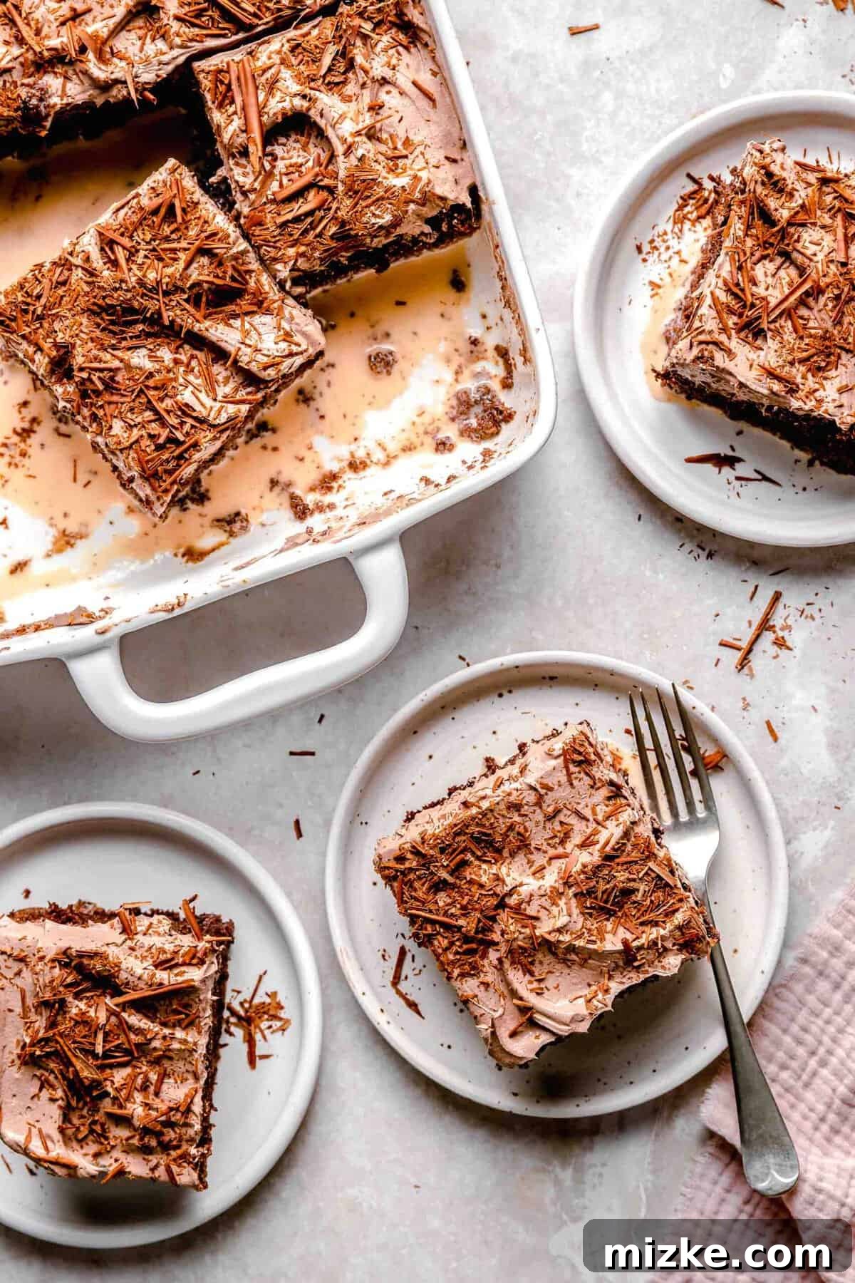 Chocolate cake is soaked in 3 kinds of milk and topped with chocolate Cool Whip and shaved chocolate in this spin on tres leches cake. A slice of moist chocolate tres leches cake on a plate with a fork, next to a full baking dish of the cake.