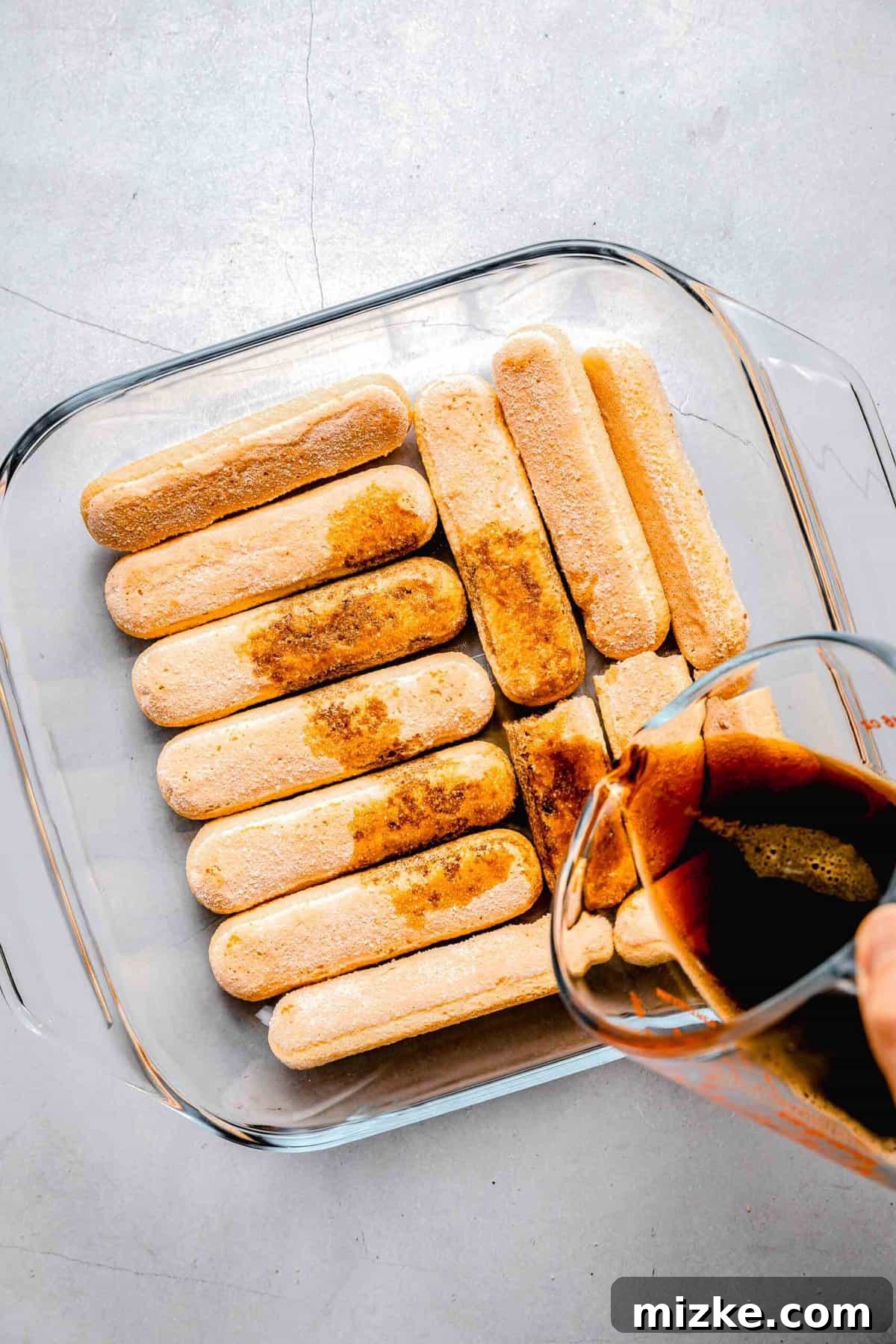 This simple tiramisu is made up of spongy, espresso-soaked ladyfingers layered with a cloudy, creamy mascarpone filling. Pouring espresso over ladyfingers in a baking gpan.