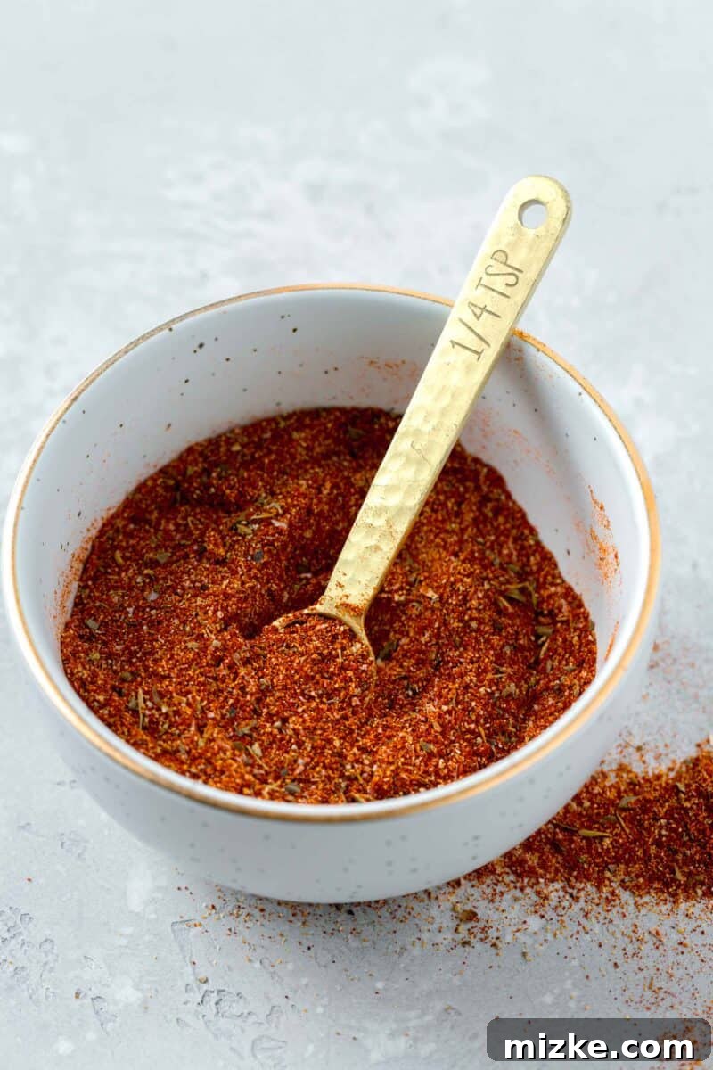 A close-up shot of homemade blackening seasoning in a small bowl, with a gold 1/4 teaspoon measuring spoon partially submerged, reflecting light. The seasoning has a rich, reddish-brown hue, and a portion is spread out on the table.