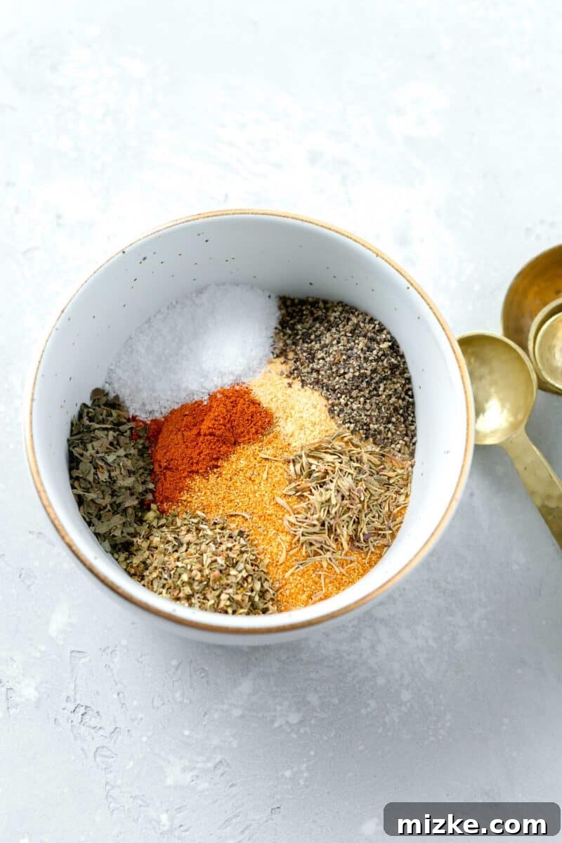 All the blackening seasoning ingredients – smoked paprika, cayenne pepper, onion powder, garlic powder, black pepper, kosher salt, dried oregano, dried basil, and dried thyme – are combined in a single white bowl with a gold rim. Gold measuring spoons are laid beside the bowl.