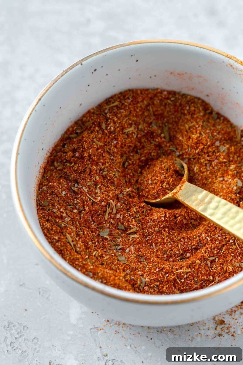 A beautifully composed shot of a glass bowl holding blackening seasoning, with a gold 1/4 teaspoon measuring spoon. The seasoning is dark red-brown, and some is artfully spilled on the table. A small glass jar with more seasoning and a wooden spoon are in the background.