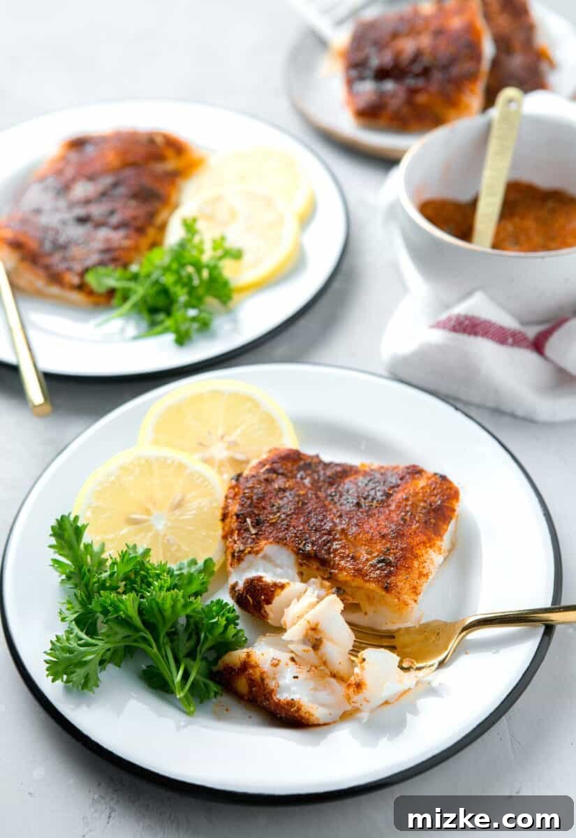 Cajun Spiced Cod Fillet 3 cod filet on a white and black rimmed plate with a gold fork piercing the flesh next to lemons and parsley