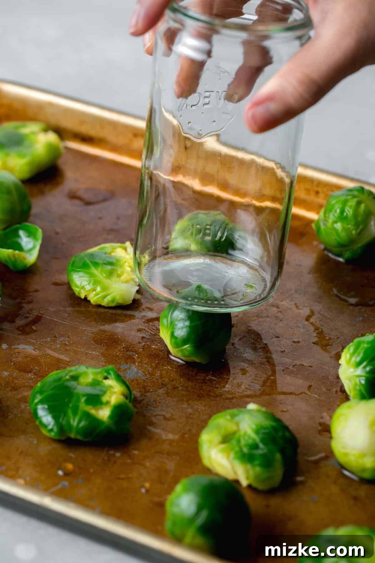 Perfectly Crisped Air Fryer Smashed Brussels Sprouts 4 Brussels sprouts on a rimmed baking sheet with a flat bottom glass being pushed on top of a Brussels sprout to smash it