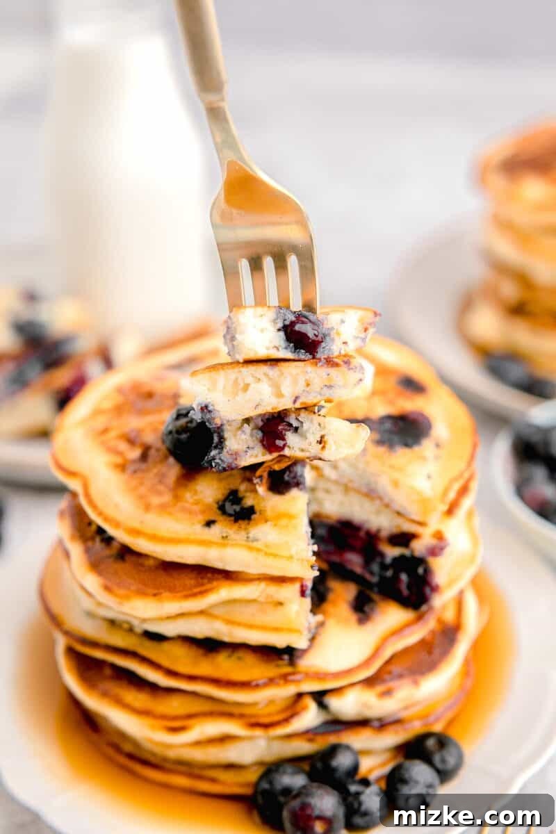 Sweet Blueberry Stacks 11 A fork is delicately piercing into a few pieces of a fluffy blueberry pancake, lifted from a tall, inviting stack, showcasing its tender interior and juicy berries.