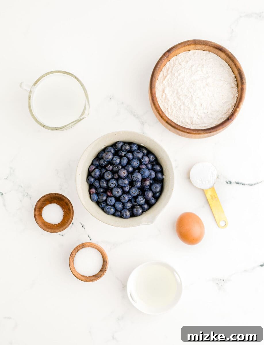 Sweet Blueberry Stacks 3 A collection of fresh ingredients laid out on a clean surface, including flour, sugar, baking powder, salt, an egg, milk, vegetable oil, and ripe blueberries, ready for making pancakes.
