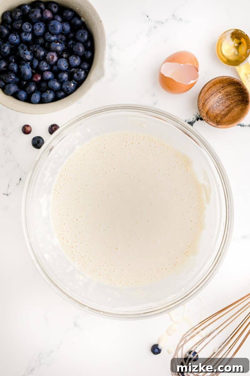 Sweet Blueberry Stacks 6 A bowl of freshly mixed, slightly lumpy pancake batter is displayed on a white surface, with a separate bowl of vibrant blueberries positioned nearby.