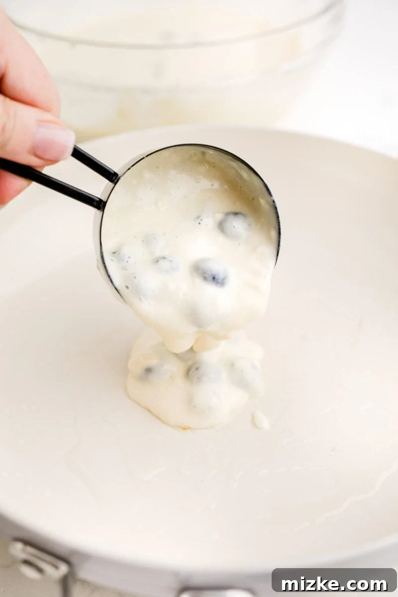 Sweet Blueberry Stacks 8 Smooth blueberry pancake batter being poured precisely from a measuring cup onto a hot, white non-stick skillet, forming a perfect circle.