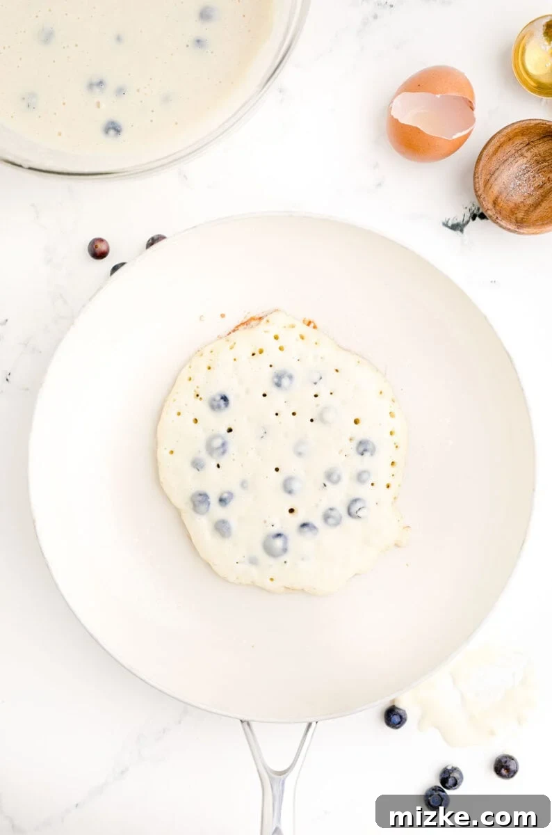 Sweet Blueberry Stacks 9 A perfectly circular pancake, studded with blueberries, cooking on a large white non-stick skillet, with a golden-brown bottom and active bubbles forming on top.