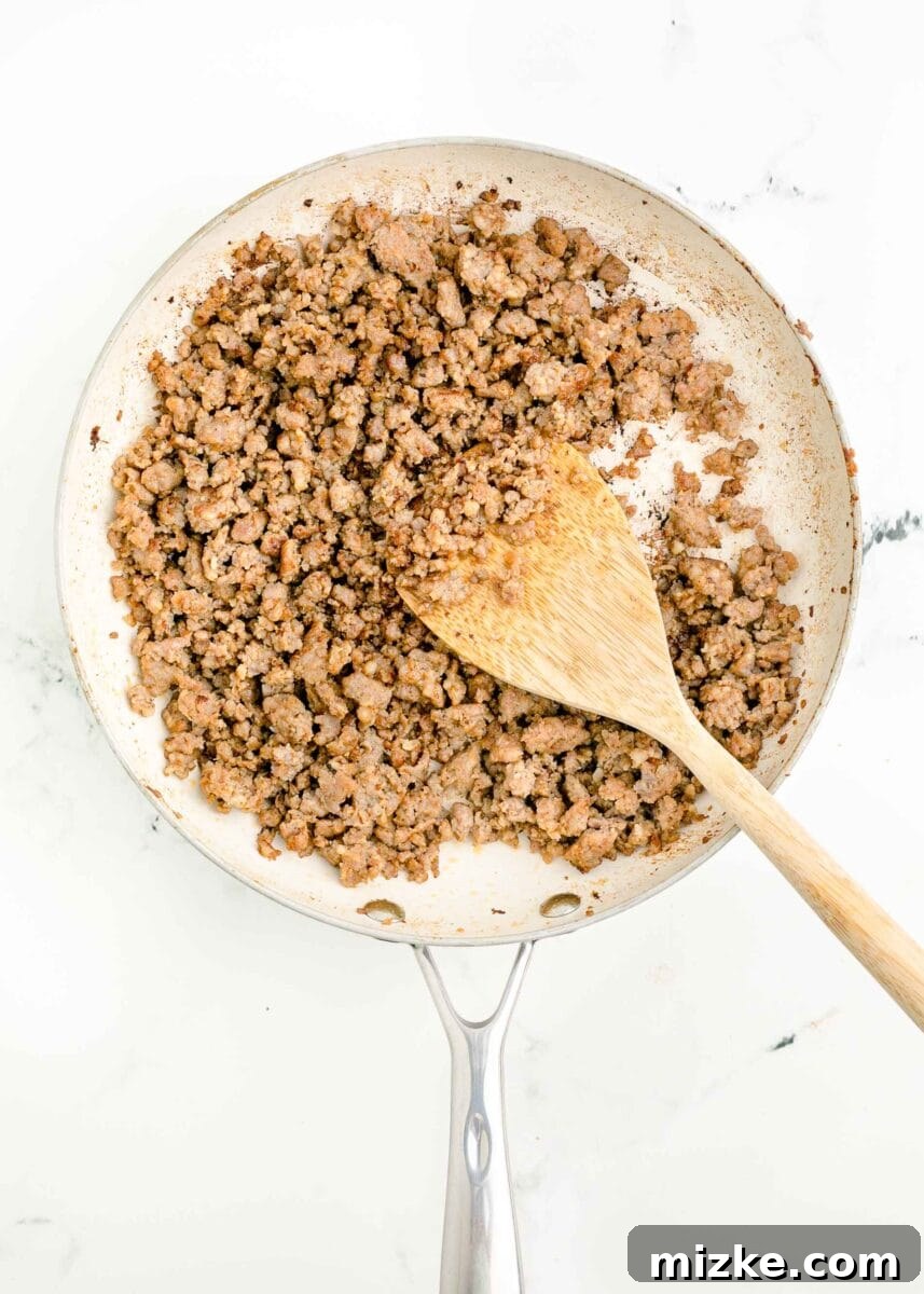 Sunrise Crunch Fold 4 A wooden spoon actively cooking and breaking apart ground breakfast sausage in a large skillet, ensuring even browning.