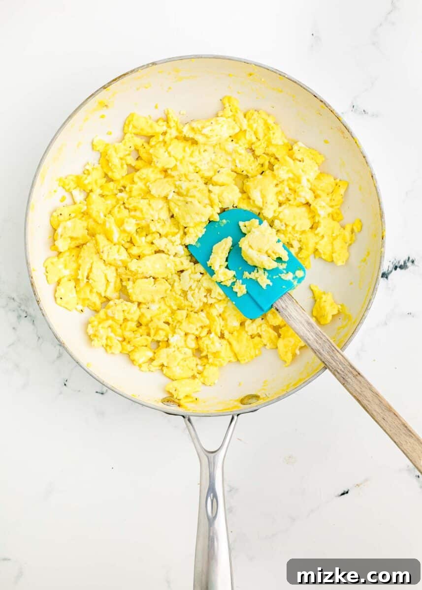 Sunrise Crunch Fold 5 Fluffy scrambled eggs are being gently cooked in a non-stick skillet using a flexible rubber spatula, ensuring a soft and even texture.