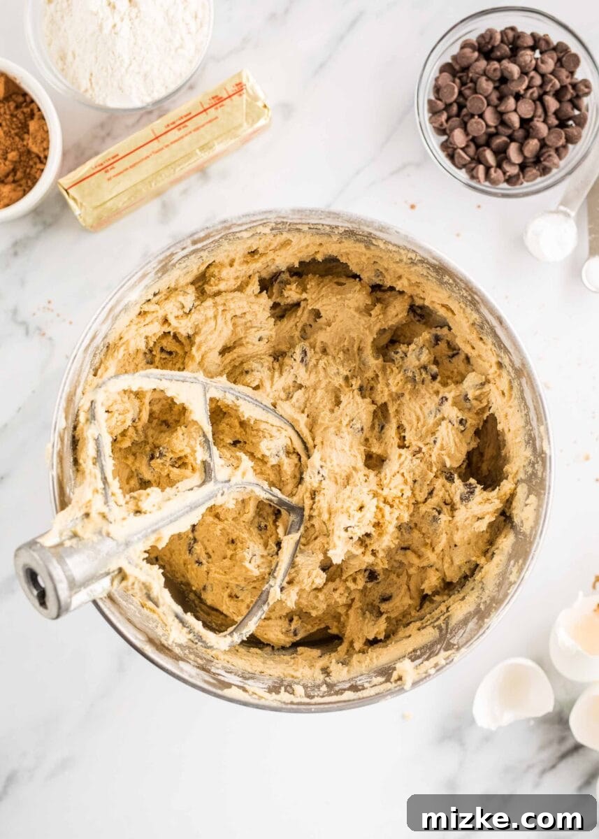 Brownie Cookie Fusion 7 Fluffy, golden-brown chocolate chip cookie dough has been expertly mixed with an electric mixer in a large bowl, showcasing its readiness for baking.