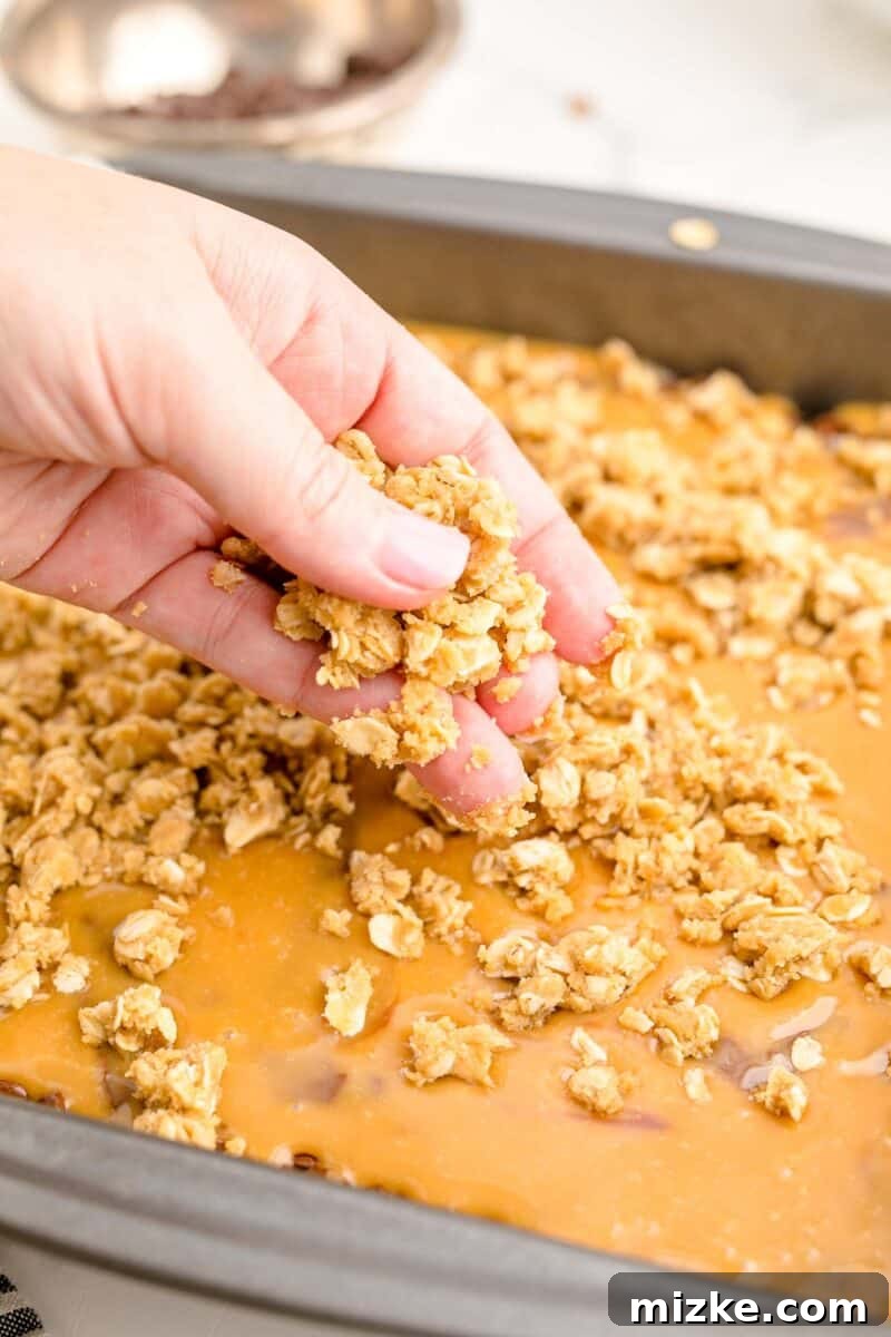 The Carmelitas Legacy 10 A hand sprinkles the remaining oat crust mixture over the caramel and chocolate layers in the baking dish.