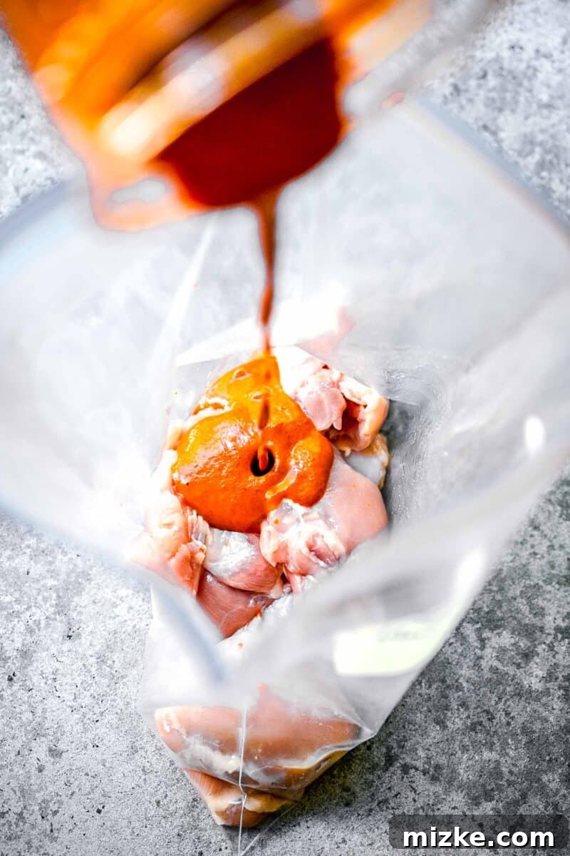 Smoky Chipotle Grilled Chicken 4 A vibrant, red-orange marinade is being poured generously on top of boneless, skinless chicken thighs inside a large plastic bag for marination.