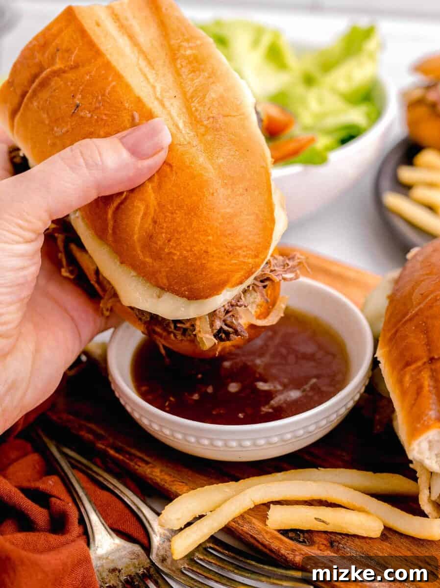 Effortless Slow Cooker French Dip Perfection 11 A delicious French Dip sandwich, with its layers of tender beef and melted cheese, is being dipped into a small bowl of rich au jus, highlighting the interactive and flavorful experience.