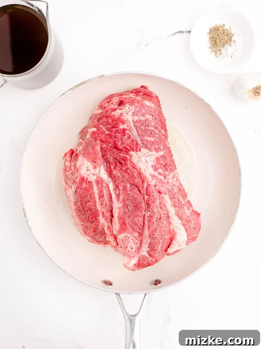 Effortless Slow Cooker French Dip Perfection 4 A raw beef roast, generously seasoned with salt and pepper, awaits searing in a pristine white skillet.