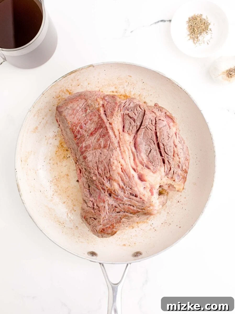 Effortless Slow Cooker French Dip Perfection 5 The beef roast is beautifully seared to a golden-brown crust on one side, still in the white skillet, hinting at the flavor development.