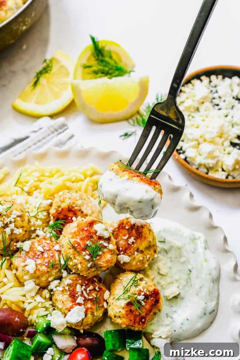 Zesty Greek Chicken Meatballs 9 A single, perfectly cooked Greek chicken meatball being dipped into a creamy white sauce, highlighting its tender texture.
