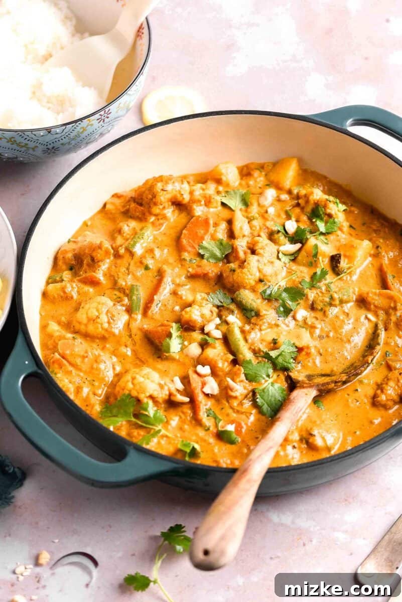 A wooden mixing spoon rests inside a large pot brimming with freshly cooked vegetable korma, showcasing its rich, creamy texture and vibrant vegetable medley.
