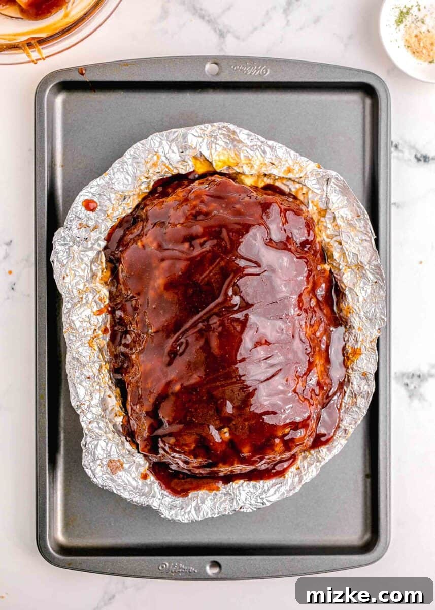 Perfect Instant Pot Meatloaf 11 A perfectly glazed meatloaf is carefully placed in its protective foil pan on a baking sheet, strategically positioned for the final broiling step to achieve a bubbly, caramelized crust.