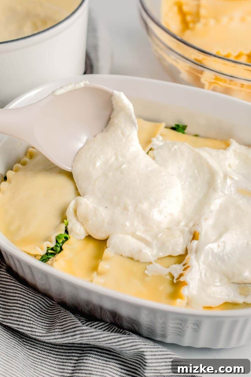 A spoon smooths a creamy layer of white sauce over the cooked lasagna noodles in a baking dish.