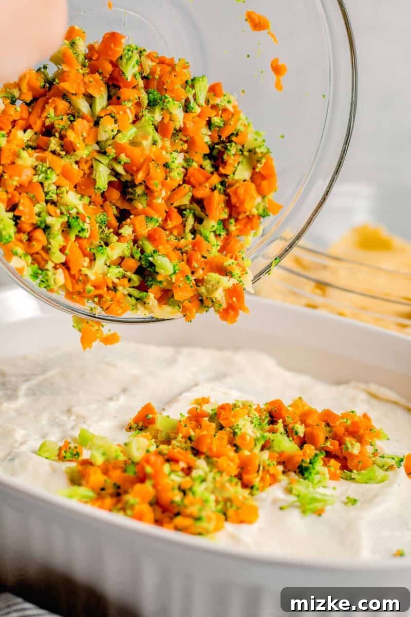 A colorful medley of vibrant chopped vegetables is being sprinkled generously on top of a creamy layer of white sauce in a lasagna dish.