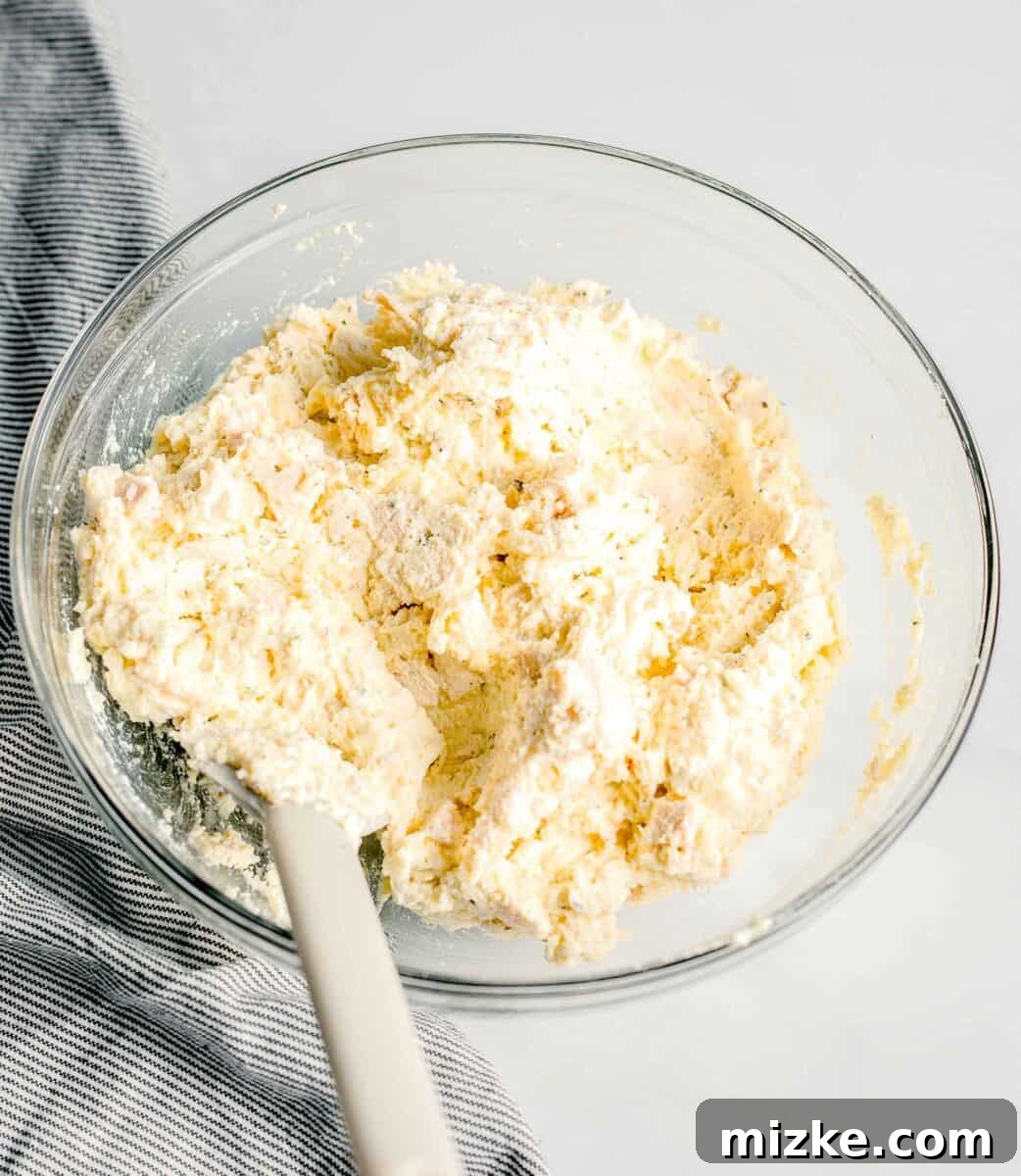A cheesy egg mixture, infused with herbs, is thoroughly combined in a glass mixing bowl, forming the base for the chicken filling.