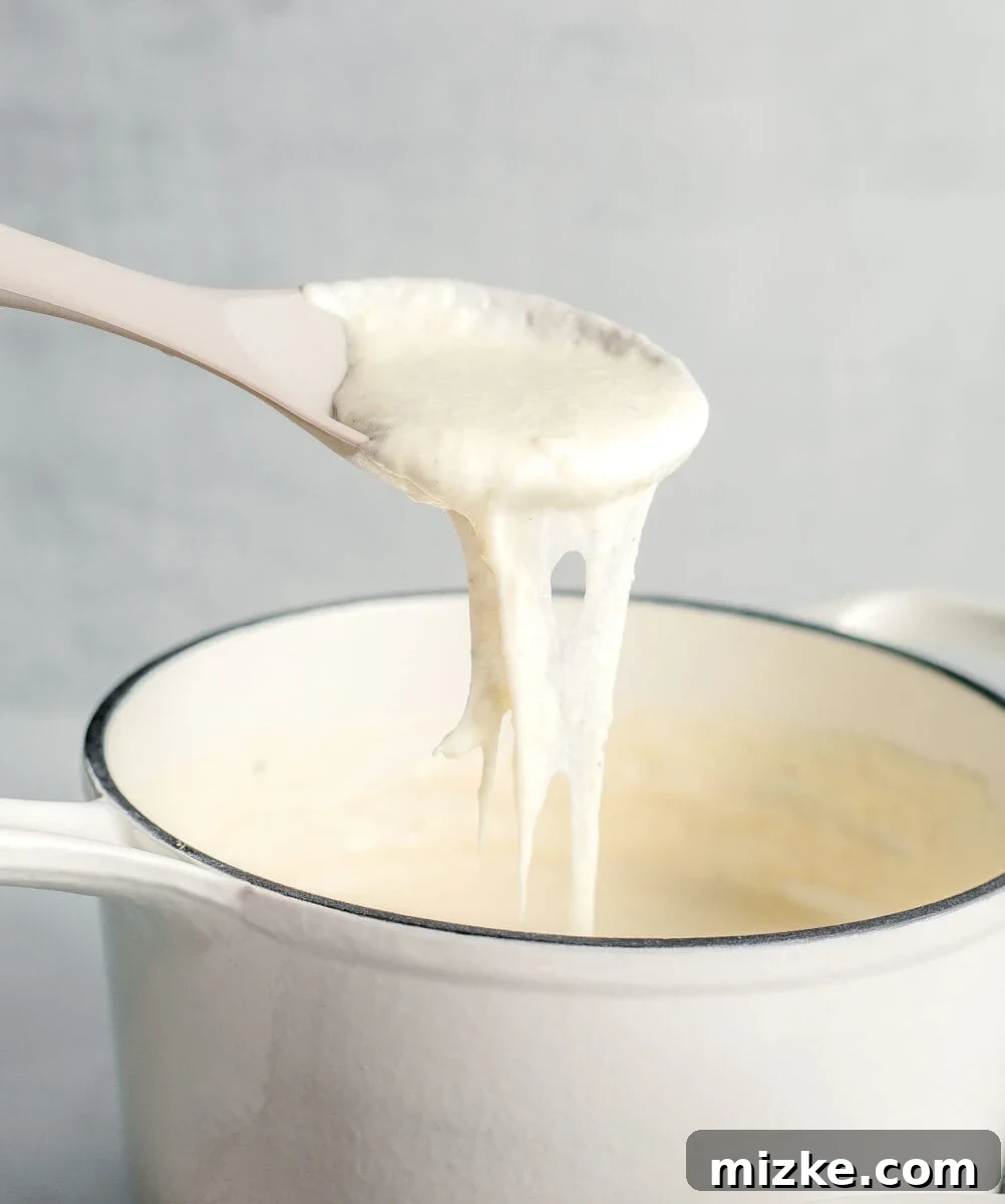 A spoon actively stirs a pan of creamy white cheese sauce, ensuring a smooth and lump-free consistency as it thickens.