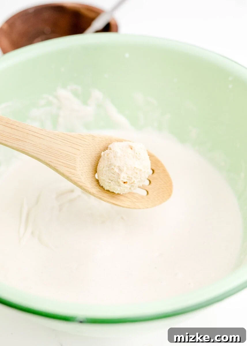 Crispy Cheesecake Poppers 7 A perfectly formed ball of creamy cheesecake mixture is gently held above a bowl of liquid batter, poised for its delicious coating.