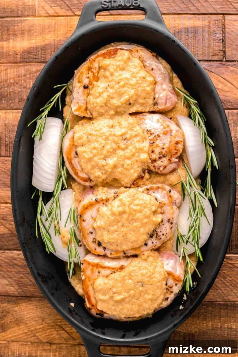 Juicy Oven-Roasted Pork Chops 8 Uncooked pork chops, surrounded by onion quarters and fresh rosemary herbs, are arranged in a black baking pot, ready for the oven.