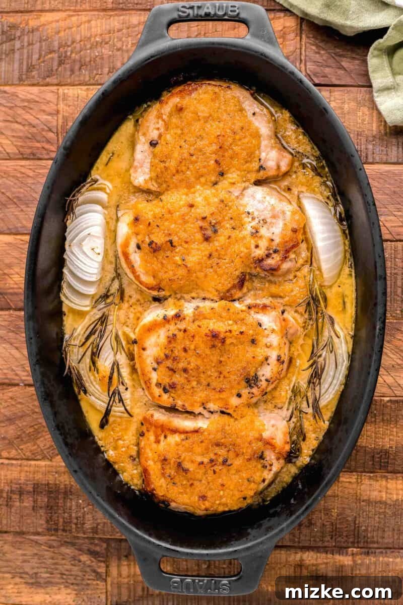 Flavorful Oven Roasted Pork Chops 9 Four beautifully coated pork chops, garnished with fresh rosemary and onion quarters, ready to be baked in a black casserole dish.