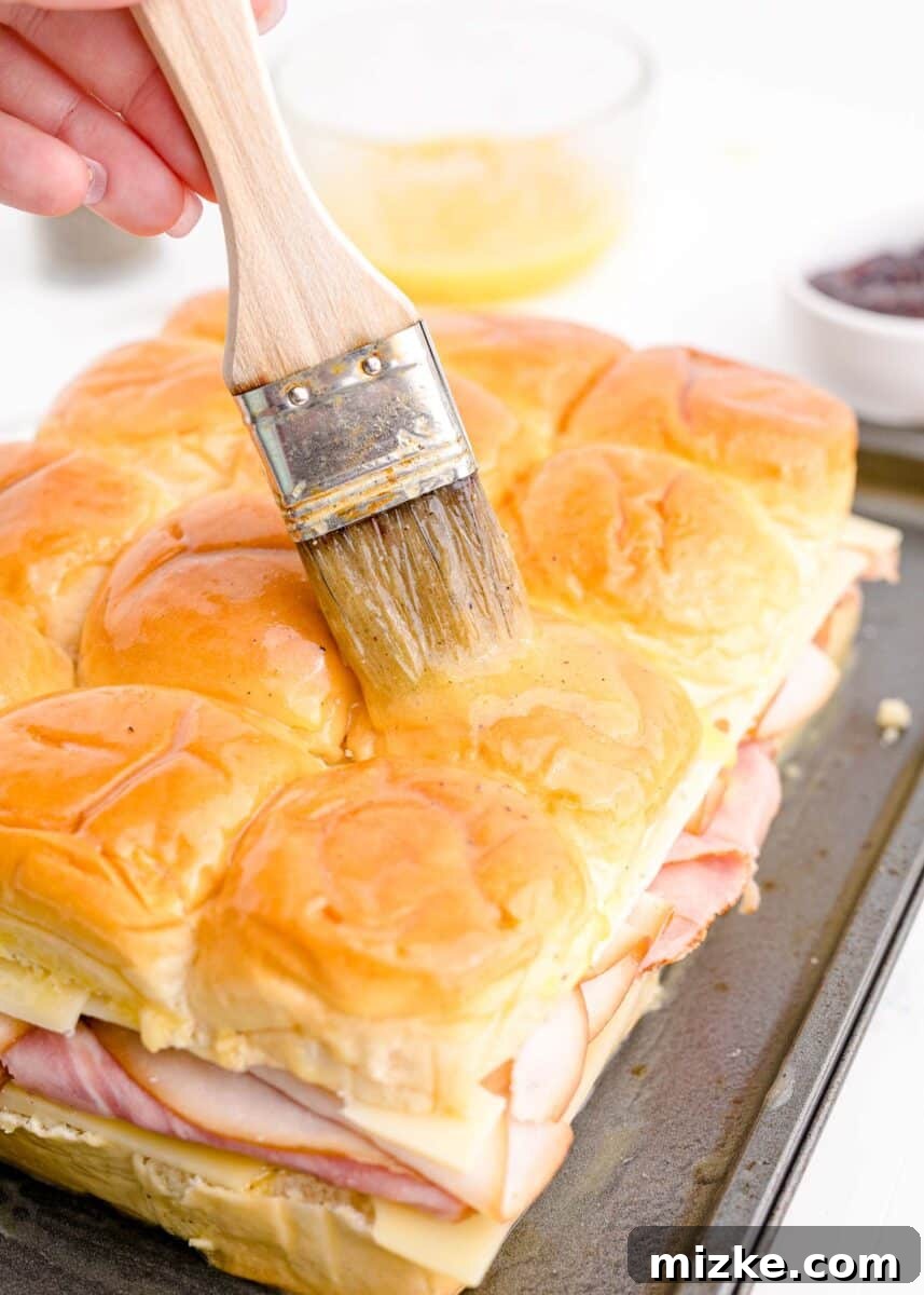 A pastry brush is meticulously applying the buttery egg mixture over the tops of the assembled sliders in a baking dish, preparing them for the oven.