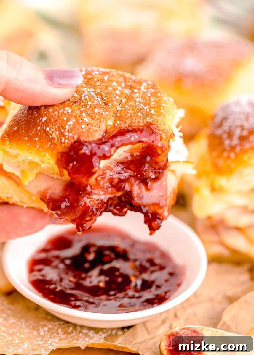 A Monte Cristo Slider is being delicately dipped into a small bowl of vibrant red jam, showcasing the classic sweet and savory pairing.