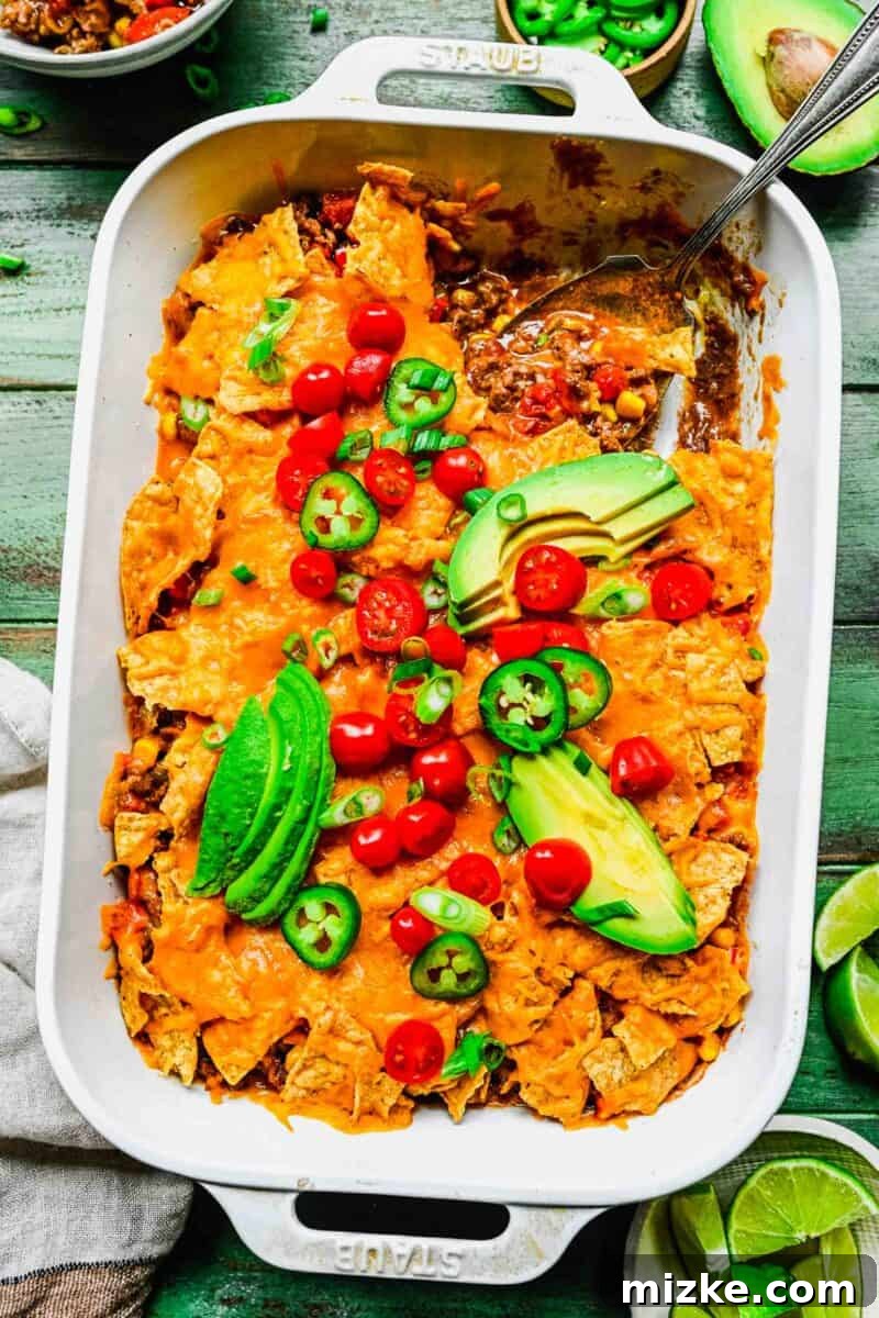 This easy taco casserole recipe is a cheesy, beefy, flavor-packed dinner idea that's ideal for crazy weeknights and parties alike! A vibrant taco casserole in a white baking dish, ready to be served. It's garnished with fresh tomatoes, creamy avocado slices, and fiery jalapeño slices on top, showcasing its rich, layered contents.