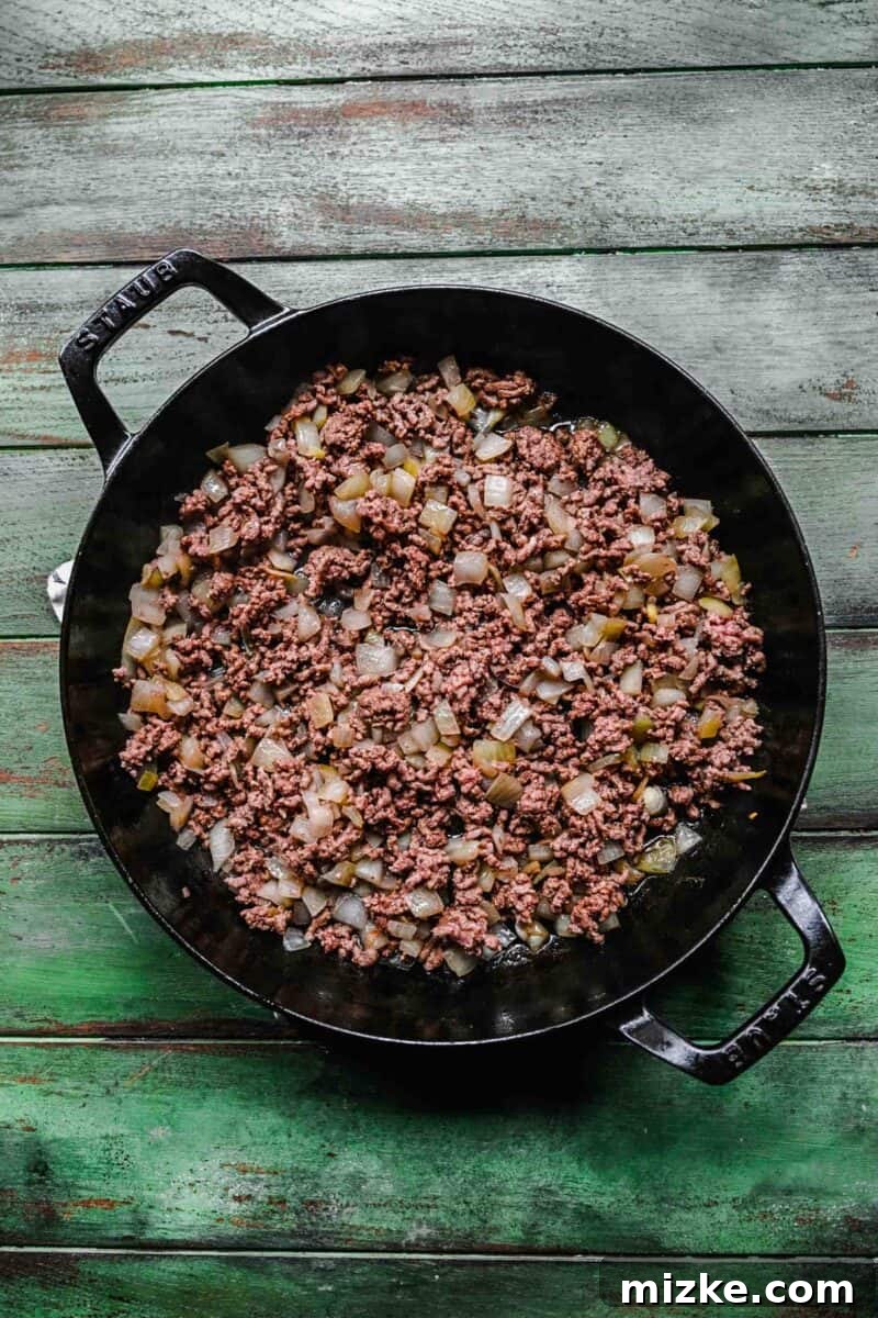 Sautéing ground beef and onions for a flavorful taco casserole. Finely diced yellow onions are sautéing with ground beef in a large skillet, creating a fragrant base for the casserole.