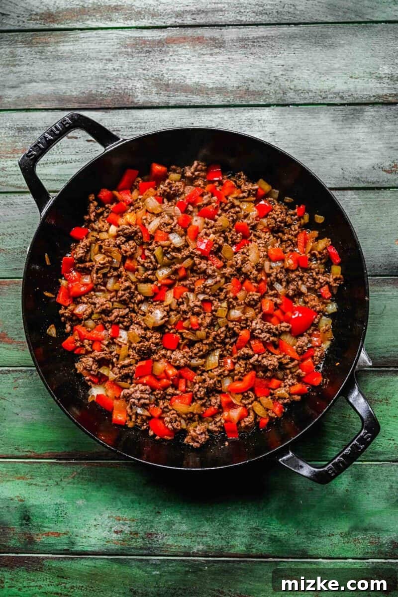 Adding bell peppers to the ground beef mixture for a colorful and tasty taco casserole filling. Vibrant red bell peppers are added to the skillet, cooking alongside the browned ground beef and onions, enriching the casserole's filling.