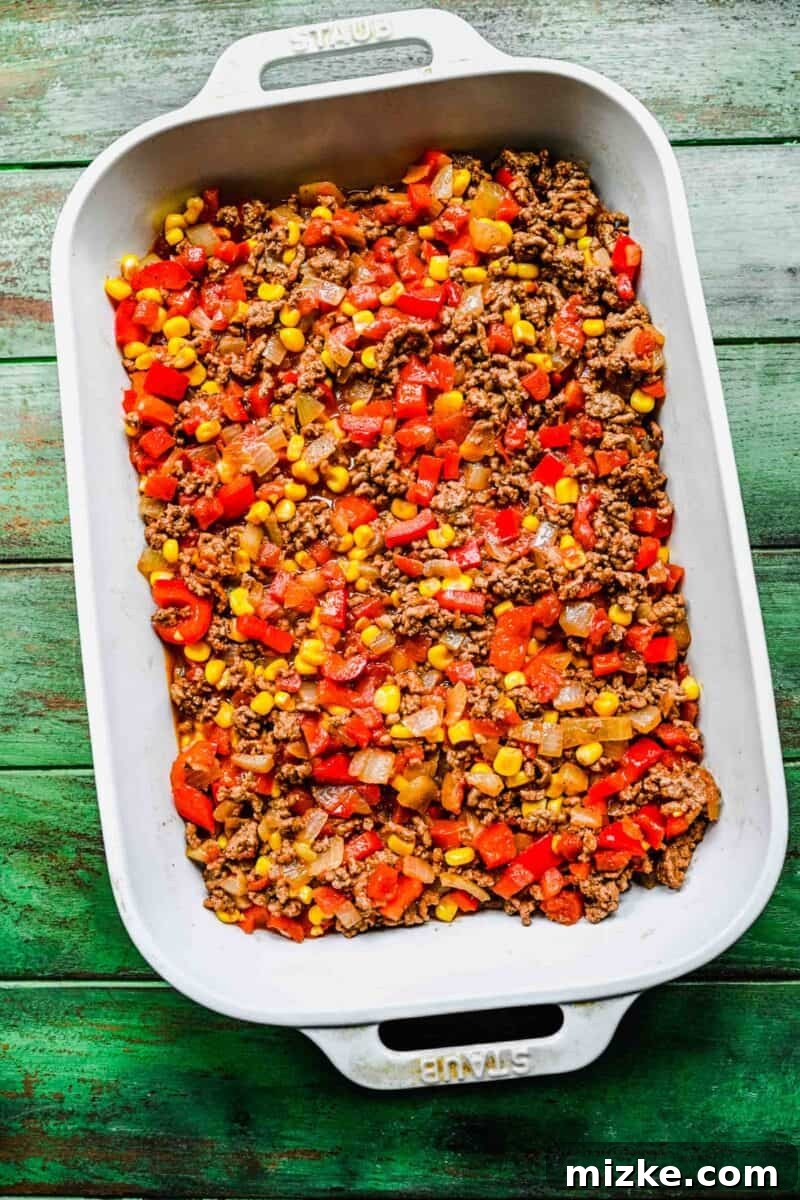 Layering the flavorful ground beef and veggie mix in the casserole dish. The rich ground beef and vegetable mixture is spread evenly as a layer in a white casserole dish, forming the heart of the taco casserole.